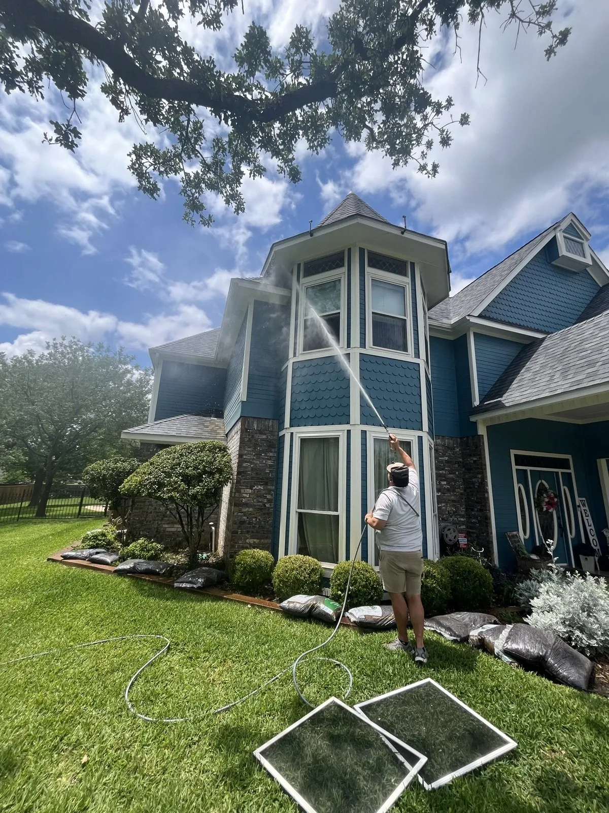 A technician cleaning the windows of a blue two-story home, screens drying on the lawn