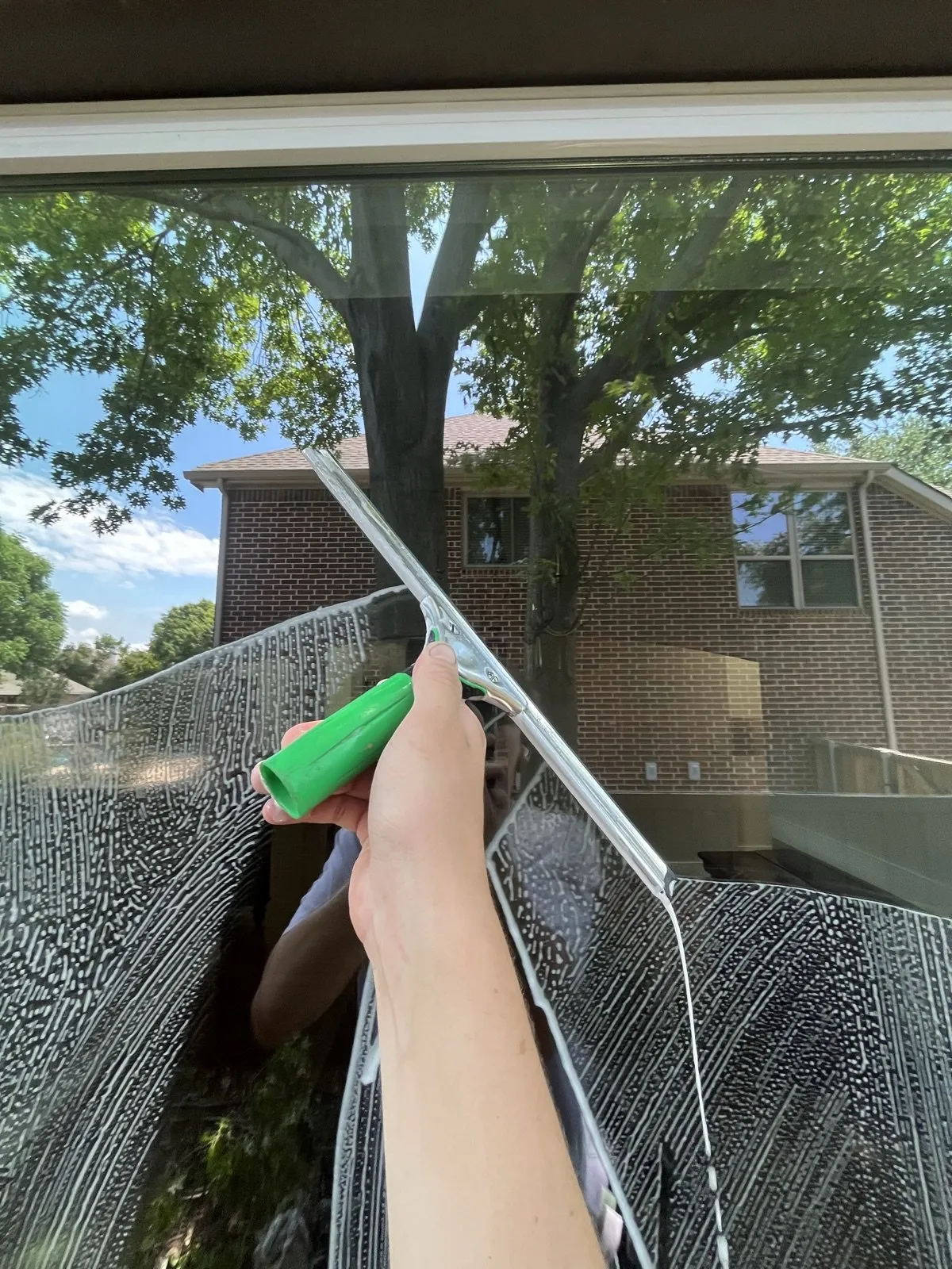 A squeegee pulling soapy water down a large window, the brick home reflected in the glass